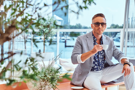 Confident Successful Man With Stylish Hair Dressed In Modern Elegant Clothes Holds Cup Of Coffee While Sitting On A Table At Outdoor Cafe Against The Background Of City Wharf.