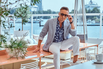 Successful stylish man dressed in modern elegant clothes sitting on table at outdoor cafe against...