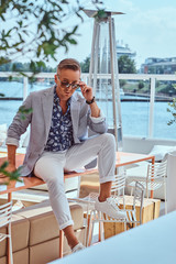Successful stylish man dressed in modern elegant clothes sitting on table at outdoor cafe against the background of city wharf.