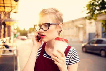 woman in glasses with coffee talking on the phone on the street evening sun