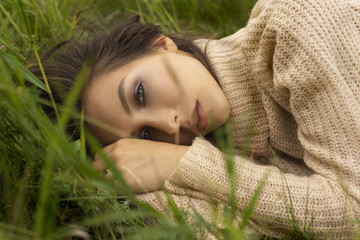 Beautiful girl wearing a beige sweater lays in a meadow among the green grass. Close-up portrait.
