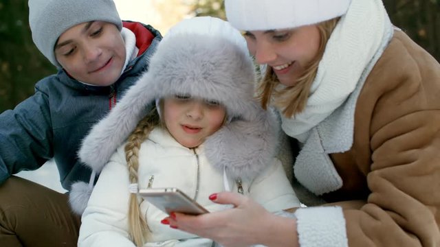Medium shot of loving young mother and two children smiling when viewing photographs on smartphone in winter forest
