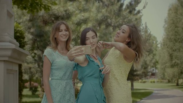 Lifestyle selfie portrait of three positive women having fun and making selfie on cellphone while resting in nature on sunny summer day. Group of happy women taking self-portrait on travel vacations.