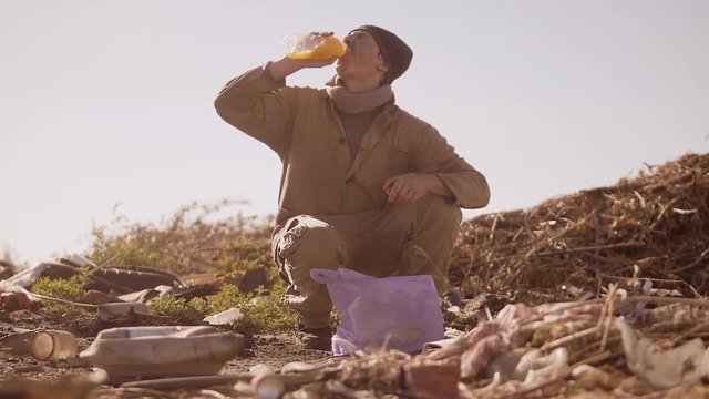 portrait of a dirty homeless hungry man in a dump drinks the missing juice in the package with walking goes looking for food slow motion video. homeless dirty man roofless person looking for food in a