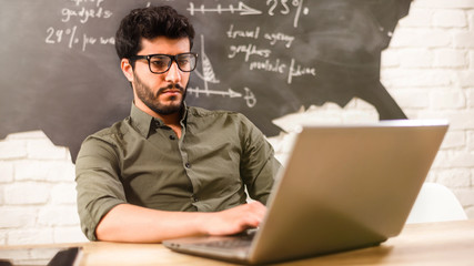 Attractive multicultural bearded businessman in glasses and green shirt working behind the laptop and writting some words in the office on the some diagrams background