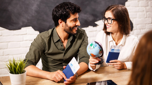Young cute couple choosing a trip with agent sitting at the travel agency office with world map on the background, they looking to each other