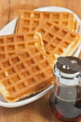 Top view, close up of four freshly baked, golden, hot waffles on an oval, white plate with a vintage, dinner type,  syrup container on a wood serving board