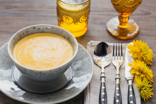 Rustic table setting with pumpkin soup, linen napkin, cutlery, ceramic plates, yellow glasses and yellow flowers