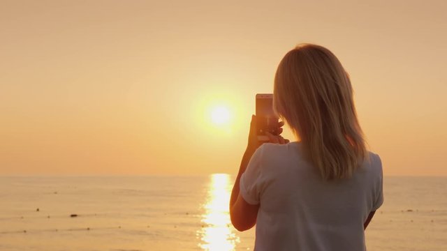 A young blonde woman is photographing on a smartphone a pink dawn by the sea. View from the back