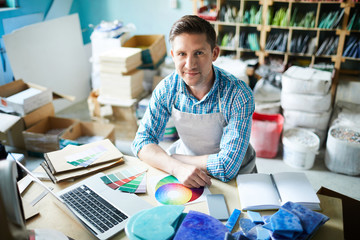 High angle portrait of modern artisan looking at camera and smiling while choosing color palette sitting at table in modern workshop, copy space