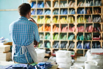 Back view portrait of modern artisan wearing apron choosing materials looking tall shelves in workshop