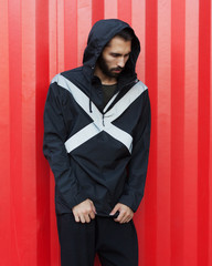 Men's youth street fashion. A bearded hipster is posing in a black jacket of new technological materials on the street, on a red background. Windbreaker