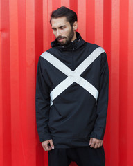 Men's youth street fashion. Windbreaker from the rain. A bearded hipster is posing in a fashionable black jacket of new technological materials on the street, on a metallic red background.