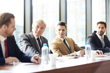 Content confident businessmen of different ages discussing company development and sitting at table in meeting room