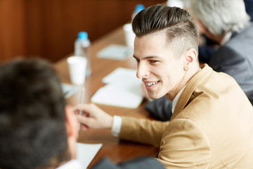 Positive handsome young business employee with fashionable hairstyle talking to colleague while sitting at table during meeting