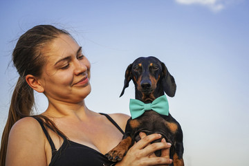 Portrait of a young beautiful girl and her dog dachshund in a blue bow, playing outdoor on the blue sky background