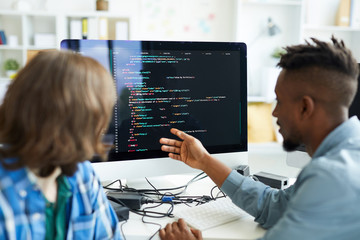 Busy young interracial colleagues analyzing computer code on computer: African man pointing at monitor and asking coworker in office