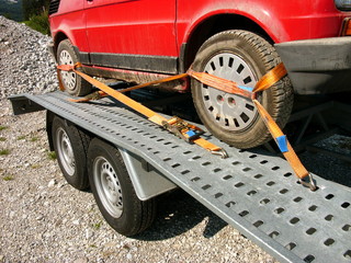 Mit Spanngurten festgezurrter roter Kleinwagen auf einem Transportanhänger auf einem Schotterweg...