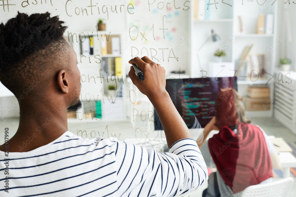 Sticker rear view of modern african-american male app developer writing code on glassy wall while working on
