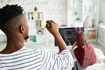 Rear view of modern African-American male app developer writing code on glassy wall while working...