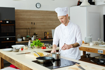 Serious concentrated handsome professional cook in white uniform making sauce and stirring it with...
