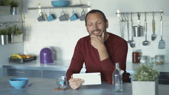 Man browsing internet on tablet in the kitchen and smiling to the camera
