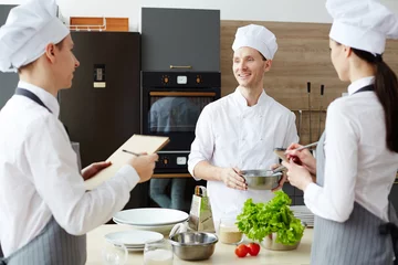 Photo sur Plexiglas Cuisinier Cheerful optimistic handsome chef in hat holding bowl with whisk while sharing cooking ideas while preparing new recipe together with colleagues at commercial kitchen