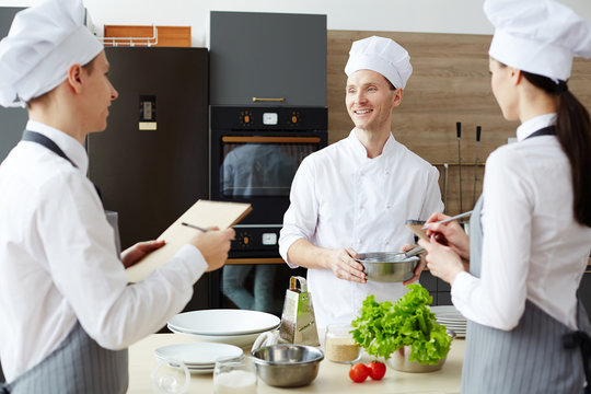 Cheerful optimistic handsome chef in hat holding bowl with whisk while sharing cooking ideas while preparing new recipe together with colleagues at commercial kitchen