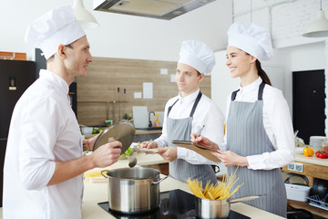 Cheerful confident interns in uniforms smiling while talking to professional chef and making notes in cooking school while listening to advises of qualified chef