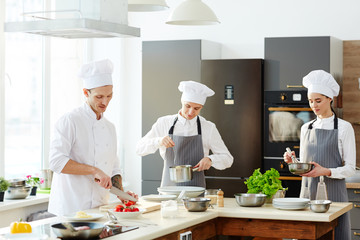 Busy chef and his cooks working at kitchen and cooking pasta at counter: chef cutting tomatoes,...