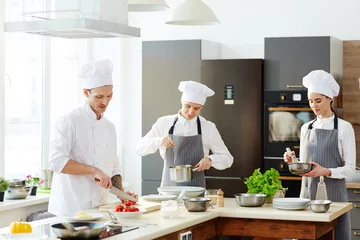 Photo sur Aluminium Cuisinier Busy chef and his cooks working at kitchen and cooking pasta at counter: chef cutting tomatoes, young man in apron checking spaghetti, serious lady whipping ingredients