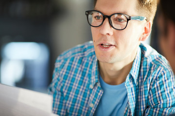 Portrait of young business professional wearing glasses and casual shirt talking to colleague while sitting at meeting table