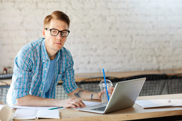 Portrait of modern young man wearing glasses and casual shirt looking at camera while working with laptop in office, copy space