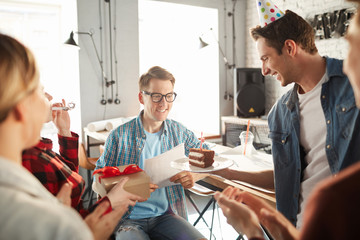 Portrait of creative young people celebrating Birthday of colleague in office, focus on man wearing glasses smiling happily receiving presents and cake, copy space
