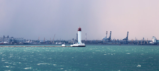 Lighthouse in the sea against the backdrop of a cargo port. Summer seascape with a white lighthouse with red top. Black Sea. Sea Port of Odessa.