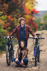 curly-haired mustachioed guy and an ore-haired girl in autumn stand with bicycles on road and show cheerful emotions.