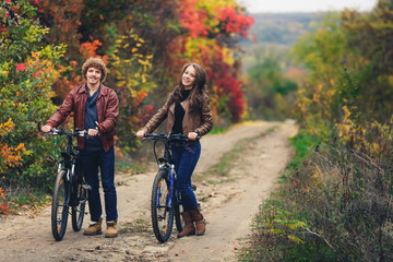Obraz premium curly-haired mustachioed guy and an ore-haired girl in autumn stand with bicycles on road and show cheerful emotions.
