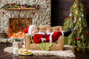 Fototapeta na wymiar A small child in a red Santa Claus costume lies on the couch in the residence, next to it there is a glass of milk and cookies on a plate. New Year card. The blond boy laughs. Christmas tree with box