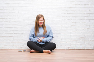 Young adult woman sitting on the floor in autumn over white brick wall with hand on stomach because indigestion, painful illness feeling unwell. Ache concept.