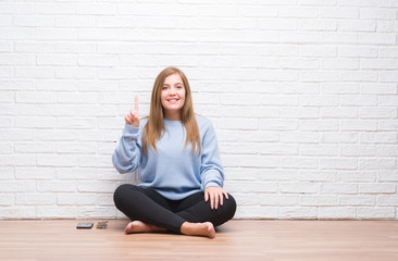 Young adult woman sitting on the floor in autumn over white brick wall showing and pointing up with finger number one while smiling confident and happy.