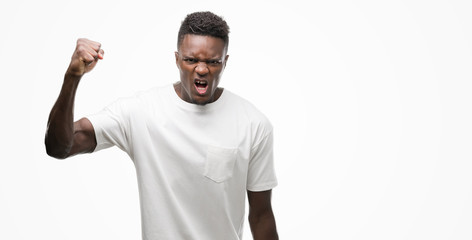 Young african american man wearing white t-shirt angry and mad raising fist frustrated and furious...