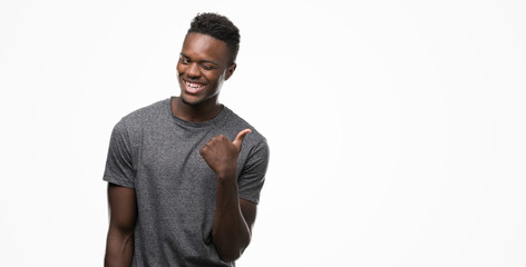 Young african american man wearing grey t-shirt smiling with happy face looking and pointing to the side with thumb up.