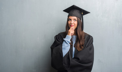 Young brunette woman over grunge grey wall wearing graduate uniform serious face thinking about...
