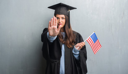 Young brunette woman over grunge grey wall wearing graduate uniform holding flag of America with...