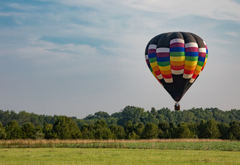 Red, Yellow, Orange, Green, and White Teardrop Shaped Hot Air Balloon Over Corn Field on Sunny Day with Trees and Cloudy Blue Sky in Background