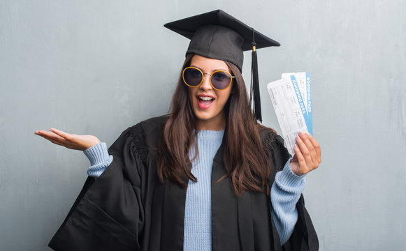 Young brunette woman over grunge grey wall wearing graduate uniform holding boarding pass very happy and excited, winner expression celebrating victory screaming with big smile and raised hands