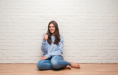 Fototapeta na wymiar Young brunette woman sitting on the floor over white brick wall happy with big smile doing ok sign, thumb up with fingers, excellent sign