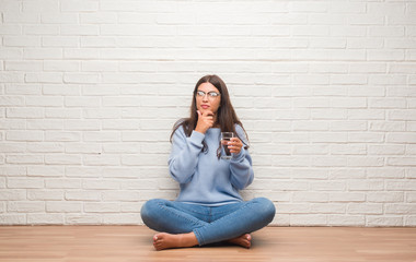 Young brunette woman sitting on the floor drinking glass of water serious face thinking about question, very confused idea
