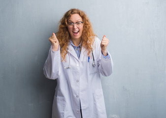 Young redhead doctor woman over grey grunge wall wearing a coat celebrating surprised and amazed...