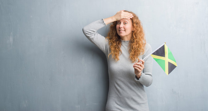 Young redhead woman over grey grunge wall holding flag of Jamaica stressed with hand on head, shocked with shame and surprise face, angry and frustrated. Fear and upset for mistake.